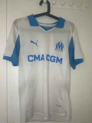 Marseille tröja Puma vit/blå - Säljer en vit och ljusblå Olympique de Marseille matchtröja från Puma. Tröjan har klubbmärke och Puma-logga på bröstet samt sponsortryck 'CMA CGM' framtill. Det står storlek M men sitter mer som S.Tillverkad i lätt och ventilerande material, perfekt för fotboll eller träning. Använd några gånger fortfarande väldigt bra skick! Tveka inte och hör av er.