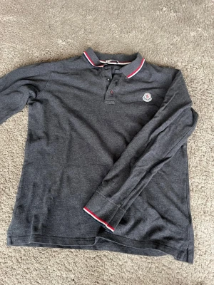 Mörkgrå Moncler långärmad piké - Snygg mörkgrå långärmad pikétröja från Moncler med klassisk krage och knappar framtill. Kragen och ärmsluten har rödvita detaljer och bröstet pryds av Monclers ikoniska logga. Perfekt för en stilren och avslappnad look. Hör av er vid frågor eller funderingar, inga defekter finns. Produkten är äkta och QR koden fungerar som den ska