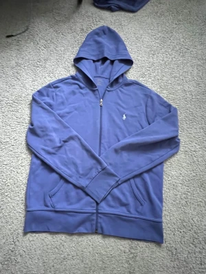 Blå hoodie med dragkedja från Polo Ralph Lauren - Snygg blå hoodie från Polo Ralph Lauren med dragkedja framtill och två fickor. Klassisk huva och det ikoniska vita Polo-märket broderat på bröstet. Tillverkad i mjukt material som känns skönt mot huden. Perfekt för en avslappnad och stilren look.