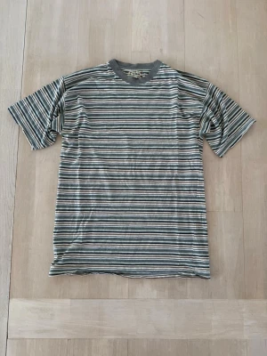Randig t-shirt med grå krage - Snygg randig t-shirt med grå ribbad krage. T-shirten har korta ärmar och tunna ränder i vitt, grått, grönt och blått. Perfekt för en avslappnad stil och passar till det mesta. Tillverkad i mjuk bomull för skön känsla.