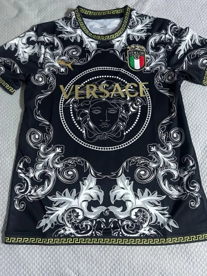 Svart  t-shirt med mönster - Unik svart t-shirt med stort Medusa-tryck och guldiga detaljer. T-shirten har korta ärmar, vita barockinspirerade mönster och ett broderat italienskt landslagsemblem på bröstet. Tillverkad i mjukt syntetmaterial och har guldfärgade Versace-loggor.