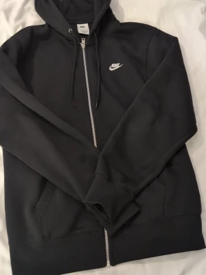 Svart zip hoodie från Nike - Klassisk svart hoodie från Nike med dragkedja framtill och vit broderad logga på bröstet. Tröjan har huva med dragsko och två fickor framtill. Perfekt för en avslappnad och sportig stil. Helt ny 