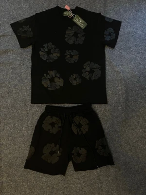  Denim Tears svart set - Svarta shorts från Denim Tears med stora blommiga mönster i mörkare nyans. Shortsen har elastisk midja med snörning för skön passform. Perfekta för en avslappnad och trendig stil.Finns i andra färger. Storlekar S och M finns