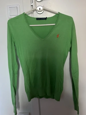 Grön stickad tröja från Ralph Lauren - Snygg grön stickad tröja från Ralph Lauren med V-ringning och lång ärm. Tröjan har den klassiska broderade loggan i orange på bröstet och är tillverkad i ett tunt, mjukt material som sitter tajt mot kroppen. Perfekt för dig som gillar stilrena plagg med en twist. Finns ett hål som jag sytt igen (bild 3)