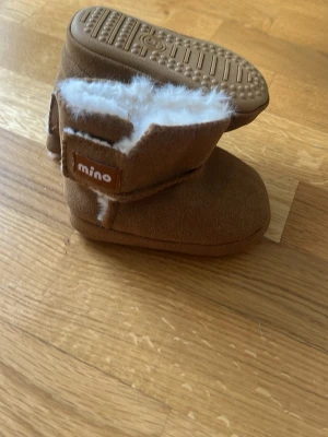 Bruna babyboots från Mino  - Säljer ett par bruna babyboots från Mino med mjukt vitt fuskpälsfoder. Bootsens utsida är i mockaimitation och de har kardborreknäppning på sidan för enkel påtagning. Perfekta för kalla dagar och riktigt gulliga på små fötter.