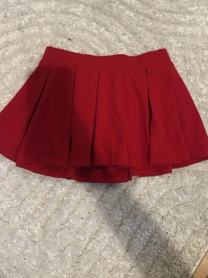 Röd plisserad skort - Snygg röd skort med plisserad design och bred linning. Kjoldelen har fina veck och under finns shorts i matchande färg. Perfekt för dig som vill ha kjol-looken men ändå känna dig bekväm. Materialet känns mjukt och följsamt.