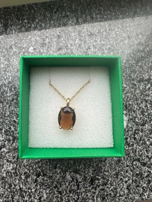 Guldhalsband med brun sten  - Helt nytt  halsband från Dyrberg/kern med en tunn guldfärgad kedja och ett stort, ovalt brunt hänge i slipad sten. Kommer i original grön ask och med vit satinpåse med Childhood-logga. Halsbandet är helt nytt och har lappen kvar på. 