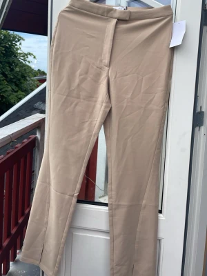 Beige kostymbyxa från NA-KD - Snygga beige kostymbyxor från NA-KD med raka ben och pressveck. Byxorna har hög midja, dold dragkedja och knapp framtill samt dekorfickor bak. Tillverkade i ett mjukt, vävt material som ger en clean look.