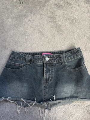 Low waist jeanskjol - Säljer ett par grå denimshorts med fransig, rå kant och klassisk femficksdesign. Shortsen har knapp och dragkedja framtill samt bälteshällor. Perfekta för dig som gillar en avslappnad och trendig stil. 