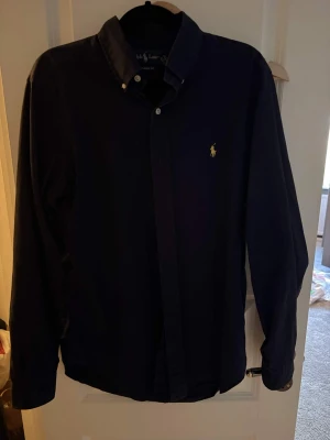 Ralph Lauren Skjorta – Mörkblå (Storlek S) - Säljer en Ralph Lauren skjorta i storlek S i bra skick. Klassisk och stilren skjorta i mörkblå färg med den ikoniska broderade Ralph Lauren-loggan på bröstet. En tidlös modell som passar lika bra till vardags som till mer uppklädda tillfällen. 🔹 Märke: Ralph Lauren 🔹 Modell: Skjorta 🔹 Färg: Mörkblå 🔹 Storlek: S 🔹 Skick: Bra skick 🔹 Passform: klassisk Väl omhändertagen och fortfarande i fint skick. Perfekt för dig som vill ha en kvalitetskjorta från Ralph Lauren till ett bra pris