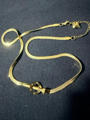 Guldigt halsband med cirkeldetalj - Snyggt guldigt halsband med en platt, ormliknande kedja och en rund cirkeldetalj framtill. Halsbandet har ett hjärtformat hänge vid låset och justerbar längd. Perfekt accessoar för att lyfta din outfit.