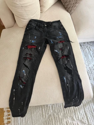 Svarta distressed jeans med röda detaljer - Säljer ett par svarta jeans med slitningar och röda mesh-detaljer under hålen. Jeansen har även vita och blå färgstänk för en extra edgy look. Femficksmodell med knappgylf och raka ben. Perfekta för dig som gillar streetwear och unik stil.