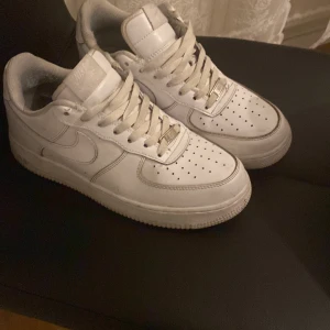 Nike Air Force 1 vita sneakers - Klassiska Nike Air Force 1 sneakers i helvitt skinn med perforerad tå och rund tå. Snygg och tidlös modell med platt sula och snörning. Perfekta för dig som gillar streetstyle och vill ha en ikonisk sneaker i garderoben.