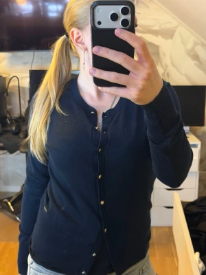 Mörkblå cardigan från Holly & Whyte - Mörkblå cardigan från Holly & Whyte med rund halsringning och knappar framtill. Långärmad modell i mjukt, tunt material med ribbade muddar vid ärmslut och nederkant. Perfekt att slänga över en t-shirt eller linne.