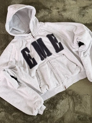 Eme Studios padded hoodie - Snygg grå hoodie med dragkedja och stora svarta bokstäver 'EME' på bröstet. Hoodien har quiltat foder på insidan för extra värme, två fickor fram och ribbade muddar. Perfekt för dig som vill ha en streetwear-look med komfort. Boxy Cropped fit.