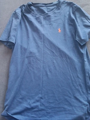 Blå t-shirt från Polo Ralph Lauren - Säljer en blå t-shirt från Polo Ralph Lauren med klassisk rosa broderad logga på bröstet. T-shirten har rund hals och korta ärmar, perfekt för en avslappnad och stilren look. Materialet är mjuk bomull som känns skönt mot huden.