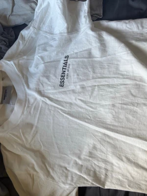 Vit Essentials T-shirt Fear of God - Vit T-shirt från Fear of God Essentials med svart tryck på bröstet. Klassisk rund halsringning och korta ärmar. T-shirten är gjord i mjuk bomull och har en clean och enkel design som är lätt att matcha.