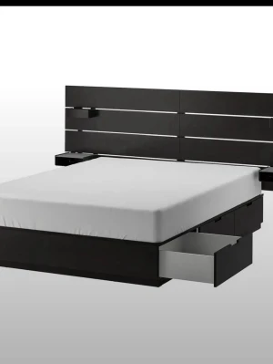 Svart sängstomme med förvaring - Modern sängstomme i svart trä med ribbad huvudgavel och praktiska lådor på sidorna för extra förvaring. Sängen har en stilren design med raka linjer och två små hyllor på huvudgaveln. Perfekt för dig som vill ha både stil och smart förvaring i sovrummet. Köpte för 3 år sen 8000kr