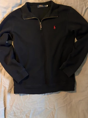 Svart halvzip från Polo Ralph Lauren - Klassisk svart tröja med halv dragkedja från Polo Ralph Lauren. Tröjan har lång ärm, hög krage och den ikoniska röda loggan broderad på bröstet. Perfekt till jeans eller chinos för en clean och stilren look.