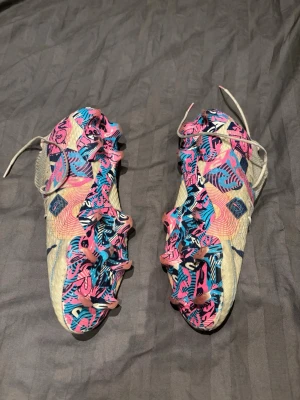 Färgglada Puma future fotbollsskor - Säljer ett par unika Puma fotbollsskor med högt skaft och snörning. Skorna har en beige bas med färgstarka detaljer i rosa, blått och lila samt grafiska mönster på sulan. Ovandelen är i textil med stickad krage för extra komfort och stöd. Storlek 45