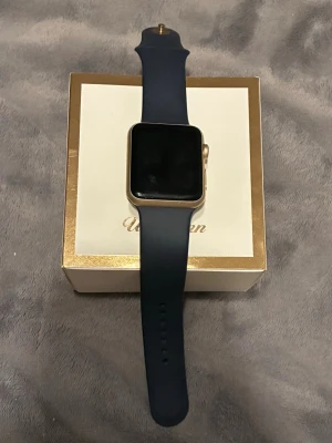 Apple Watch Series 2 42mm guld - Snygg Apple Watch Series 2 med 42mm boett i guldaluminium och svart silikonarmband. Klockan har en fyrkantig skärm och pulsmätare på baksidan. Batteriet är urladdat. Mycket bra skick som ny. 