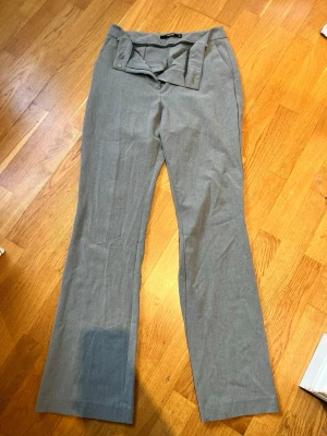 Grå kostymbyxor med bootcut - Snygga grå kostymbyxor från H&M med bootcut och hög midja. Byxorna har bred linning med knappdetalj och bälteshällor. Tillverkade i ett tunt, vävt material som ger en dressad look. Perfekta för en stilren outfit.