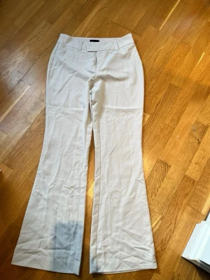 Vida beige kostymbyxor - Snygga beige kostymbyxor med utsvängda ben och hög midja. Byxorna har bälteshällor och diskreta fickor bak. Perfekta för en clean och stilren look. Materialet känns mjukt och har en lätt stretch för extra komfort.