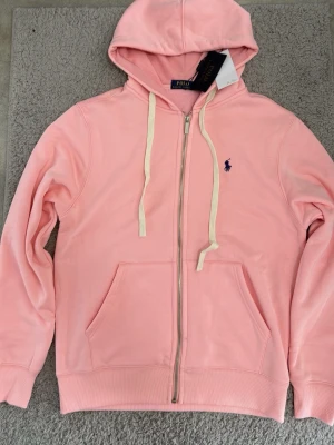 Rosa hoodie från Polo Ralph Lauren - Snygg ljusrosa hoodie från Polo Ralph Lauren med dragkedja, vit dragsko och klassisk blå logga på bröstet. Tillverkad i mjukt bomullsmaterial och har känguruficka framtill. Perfekt för en avslappnad och trendig look.