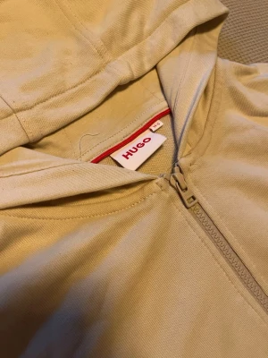 Beige hoodie med dragkedja från Hugo - Snygg beige hoodie från Hugo med dragkedja framtill och två fickor. Tvättar och stryker den innan ni går den! Knappt använd med, fråga om det är något va inte rädda!