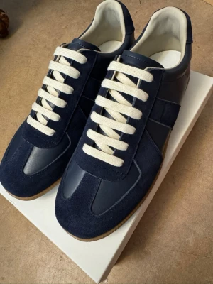  Maison Margiela Gats - Snygga marinblå sneakers från Maison Margiela med ovandel i mocka och läder, vita platta skosnören och klassisk gummisula. Stilren design med diskreta paneler och låg silhuett som passar till många olika outfits. Helt nya och oanvända.