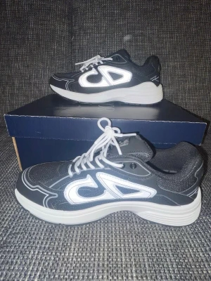 Svarta sneakers från Christian Dior - Snygga svarta sneakers från Christian Dior med vita detaljer och reflekterande CD-logga på sidan. Skorna har en chunky vit sula och är tillverkade i mesh och syntetmaterial. Perfekta för dig som vill ha en trendig och sportig look.