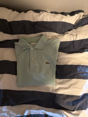 Ljusgrön pikétröja från Lacoste - Snygg ljusgrön pikétröja från Lacoste med klassisk krage och två knappar framtill. Tröjan har korta ärmar och den ikoniska krokodilloggan på bröstet. Perfekt för en clean och stilren look.