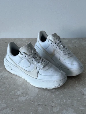 Nike Air Force 1 plt.af.orm | EU 39  - Nike Air Force 1 sneakers i helvitt läder med klassisk swoosh på sidan och AIR-detalj på sulan. Tjock sula och perforerad tå för extra ventilation. Skorna har låga skaft och vita skosnören. Perfekt för en clean och stilren look.