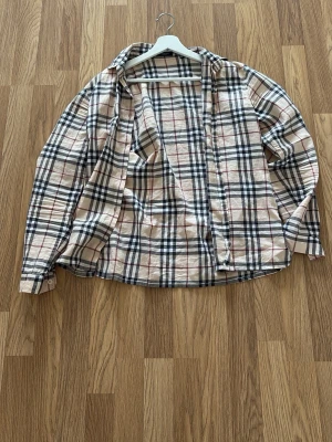 Beige rutig skjorta från Burberry - Klassisk beige rutig skjorta från Burberry med långa ärmar och knappar framtill. Skjortan har det ikoniska rutmönstret i svart, vitt och rött. Perfekt för dig som gillar tidlös och trendig stil.