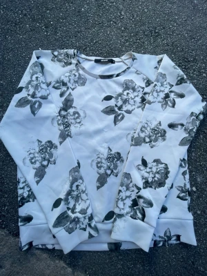 Vit sweatshirt med blommigt tryck - Säljer en vit sweatshirt från Bik Bok med svartgrått blommigt tryck över hela tröjan. Tröjan har rund halsringning och långa ärmar. Perfekt för dig som gillar en stilren men ändå unik look. Materialet känns mjukt och bekvämt mot huden.