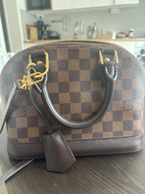 Louis Vuitton Damier Alma BB handväska - Säljer en ikonisk Louis Vuitton Alma BB handväska i brunt Damier Ebene-mönster. Väskan har rundad form, dubbla handtag i mörkbrunt läder, guldfärgade metalldetaljer och avtagbar axelrem. Klassisk och tidlös design med Louis Vuitton Paris ingraverat på läderdetaljen. Den är köpt av min kille på NK för fyra år sedan så jag har inte kvar kvittot men det går att verifierar äktheten på annat sätt om man vill. Pris kan diskuteras. 