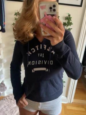 Mörkblå hoodie från Abercrombie & Fitch - Mörkblå hoodie från Abercrombie & Fitch med tryck på bröstet där det står 'A&FITCH 1st DIVISION'. Hoodien har huva med dragsko och en stor magficka. Perfekt för en avslappnad och cool stil.