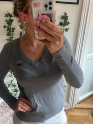 Grå stickad tröja från Hollister - Snygg grå stickad tröja från Hollister med V-ringning och diskret logga i rött och vitt på bröstet. Tröjan har långa ärmar och ribbade muddar vid ärmslut och nederkant. Perfekt till jeans eller kjol för en avslappnad look.