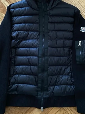 Svart jacka med huva från Moncler - Snygg svart dunväst från Moncler med huva och dragkedja framtill. Västen har quiltad framsida i glansigt material och stickade partier på sidor och ärmar. Moncler-logga på ärmen och praktisk ficka med dragkedja. Perfekt för lager-på-lager-stil.