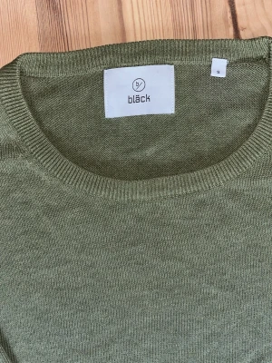 Olivgrön stickad tröja från Bläck - Snygg olivgrön stickad tröja från Bläck med rund halsringning och ribbade muddar vid ärmslut och nederkant. Perfekt för lager-på-lager och enkel att matcha med jeans eller chinos. Tunn och mjuk kvalitet som känns skön mot huden.