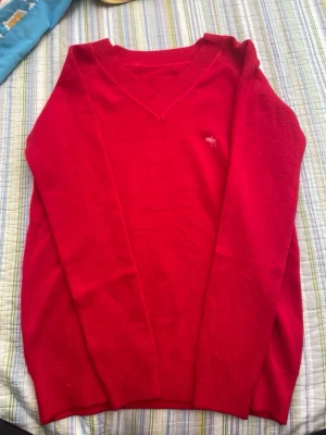 Röd stickad tröja Abercrombie & Fitch - Snygg röd stickad tröja från Abercrombie & Fitch med V-ringning och ribbade muddar. Tröjan har en liten broderad älg-logga på bröstet och är tillverkad i mjukt material. Perfekt för lager på lager och enkel att matcha.