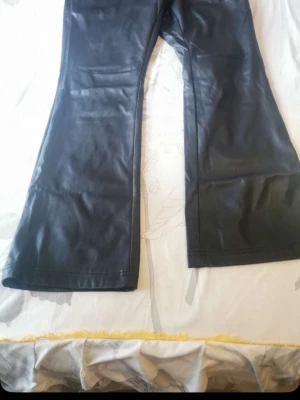 Svarta utsvängda skinnbyxor - Säljer ett par svarta skinnbyxor med utsvängda ben. Byxorna har en slät finish och är tillverkade i skinnimitation. Perfekta för dig som vill ha en edgy och trendig look.