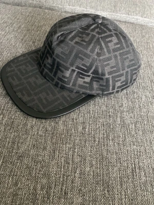 Svart keps från Fendi med logomönster - Säljer en svart keps från Fendi med det klassiska FF-logomönstret över hela kepsen. Skärmen är i svart material och kepsen har justerbart band bak med metallogo där det står 'Fendi Roma'. Perfekt för dig som gillar exklusiva accessoarer.