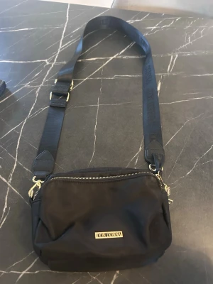 Svart axelväska från Don Donna - Snygg svart axelväska från Don Donna med justerbar rem och guldfärgade detaljer. Väskan har en dragkedja upptill och en stilren, rektangulär form. Perfekt för att bära dina viktigaste grejer och passar till många olika outfits.