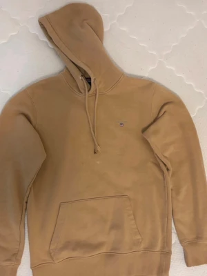 Beige hoodie från Gant  - Snygg beige hoodie från Tommy Hilfiger med klassisk huva och dragsko. Liten logga på bröstet och stor känguruficka framtill. Perfekt för en avslappnad och trendig stil. Mjuk bomullsblandning som känns skön mot huden.
