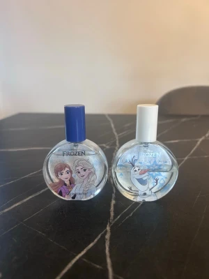 Frozen parfym duo med blå och vit kork - Två runda parfymflaskor från Disney Frozen, en med blå kork och motiv av Anna och Elsa, och en med vit kork och motiv av Olaf. Flaskorna är transparenta med snöflingor och har en lekfull design som passar fans av filmen.