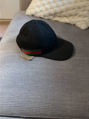 Svart keps från Gucci med GG-mönster - Snygg svart keps från Gucci med diskret GG-monogram över hela tyget. Kepsen har klassisk form, böjd skärm och justerbar rem bak. På sidan finns Guccis ikoniska röd-gröna rand. Materialet är canvas med detaljer i läder.