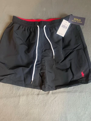 Svarta badbyxor från Polo Ralph Lauren - Snygga svarta badbyxor från Polo Ralph Lauren med vit dragsko och röd logga broderad på benet. Insidan har ett rött meshfoder och midjan är elastisk för skön passform. Klassisk design med sidofickor, perfekta för stranden eller poolen.