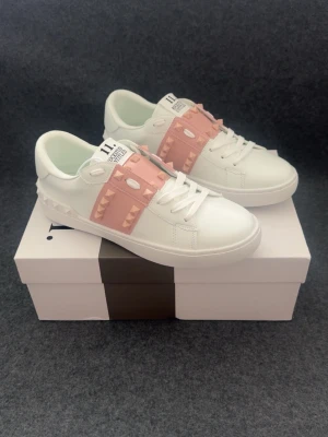 Valentino open vita sneakers med rosa detaljer - Snygga vita sneakers från Valentino open med breda rosa band och rosafärgade nitar över vristen. Skorna har en chunky vit sula med mönstrad undersida och klassisk låg profil. Tillverkade i slätt läder och har logga på plösen.