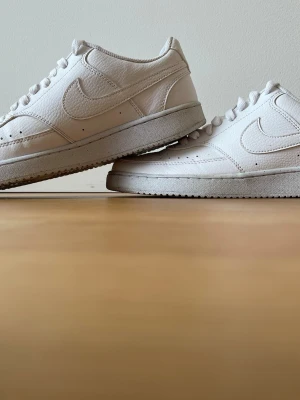 Vita Nike sneakers i läder - Stilrena vita sneakers från Nike i lågt modellutförande. Skorna är tillverkade i slittåligt läder med klassisk swoosh på sidan och perforerade detaljer vid tån. Tjock vit sula och matchande vita skosnören ger en clean look. Använda fåtal gånger. Pris kan diskuteras.